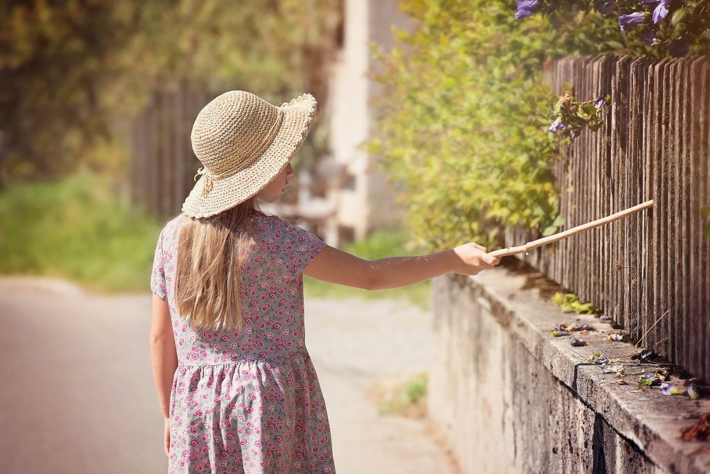 Niña rubia con sombrero de paja y vestido de flores pasea por pueblo y golpea una verja con un palo de madera