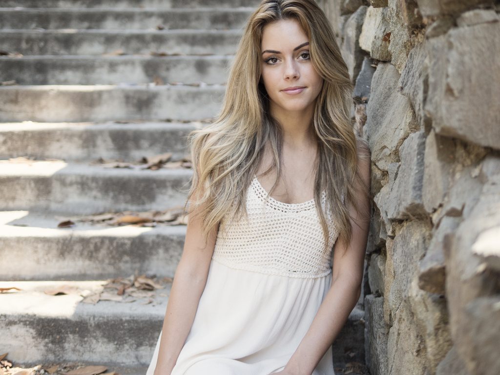 Joven Rubia con vestido blanco sentada sobre escaleras de piedra y apoyada en pared también de piedra