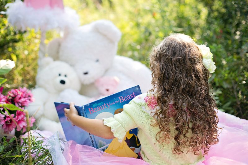 Niña leyendo "la Bella y la Bestia" en Inglés, sentada en el jardín junto a tres osos de peluche