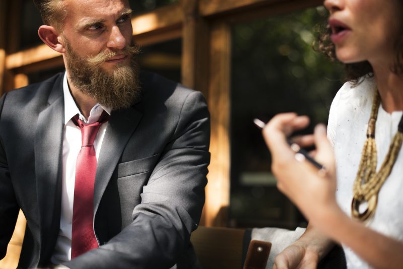 Hombre trajeado con barba y bigote escucha lo que le dice la mujer sentada a su lado