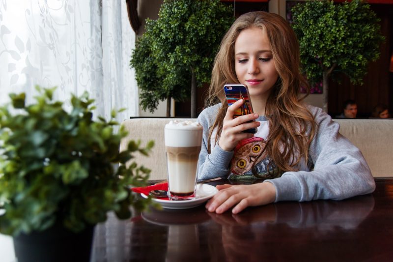 Adolescente sentada frente a un capuccino en una cafetería, mira el móvil y sonríe
