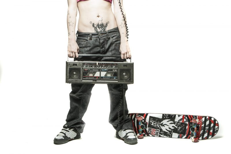 Torso de persona joven tatuada, sujetando un radiocasette y con un monopatín a su lado.