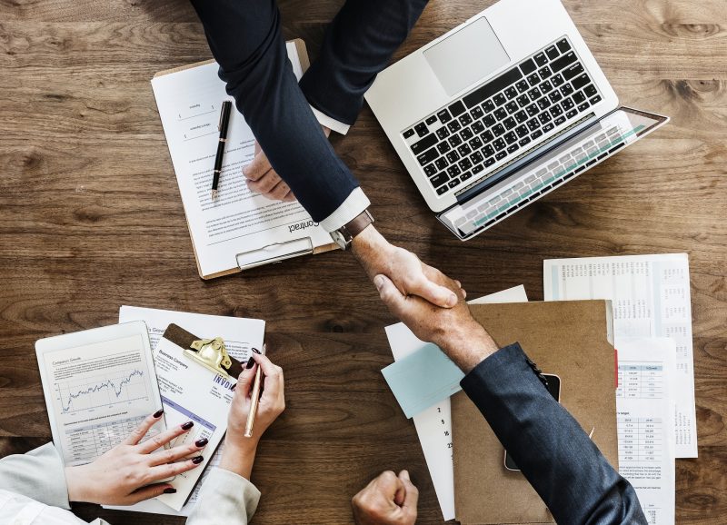 Reunión de negocios: dos miembros de un equipo se dan un apretón de manos como cierre de un contrato, mientras una tercera persona toma notas.
