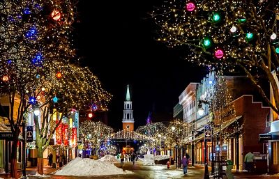 Escena navideña: ciudad con calles y árboles iluminados, nieve amontonada sobre la acera y personas paseando.