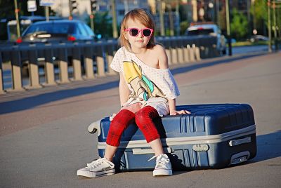 Niña con gafas de sol sentada encima de su maleta de viaje.